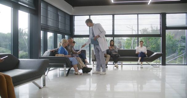 Diverse people sit on sofas in clinic lobby area, wait for appointment with doctor. Doctor speaks with elderly couple about medical test results. Waiting area in modern medical center. Healthcare. - Photo, Image