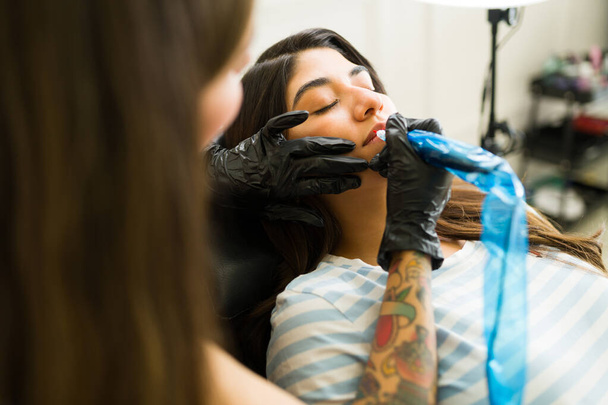Μεξικανική νεαρή γυναίκα βάζοντας σε μόνιμη χρωστική ουσία χειλιών και κάνοντας μια υπηρεσία τατουάζ χειλιών στο σαλόνι ομορφιάς - Φωτογραφία, εικόνα