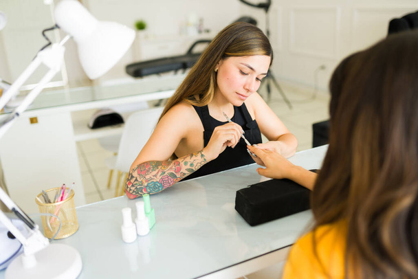 Hermosa joven que trabaja en el salón de belleza de uñas pintando las uñas para un cliente mientras hace un servicio de manicura - Foto, imagen