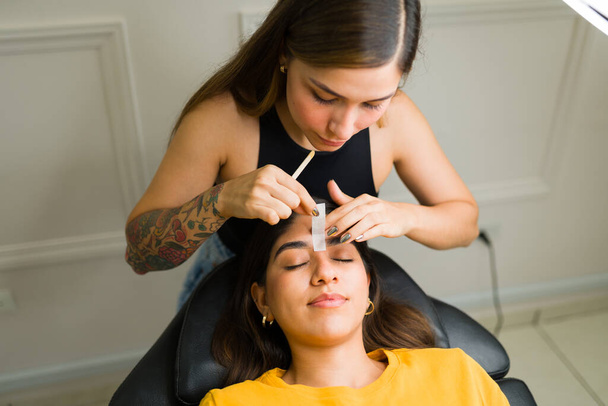 Επαγγελματική cosmetologist χρησιμοποιώντας ταινίες κερί για την απομάκρυνση των μαλλιών του προσώπου και να κάνει ένα νέο σχέδιο φρύδι για ένα χαλαρό πελάτη - Φωτογραφία, εικόνα
