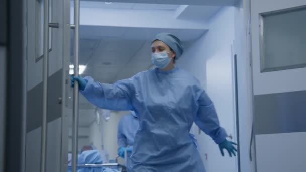 Professionele chirurgen brengen patiënten van chirurgie naar ziekenhuiskamer in de gang van het medisch centrum. Jonge vrouwelijke arts opent deuren van operatieblok voor collega 's. Patiënt ligt op brancard na operatie. - Video