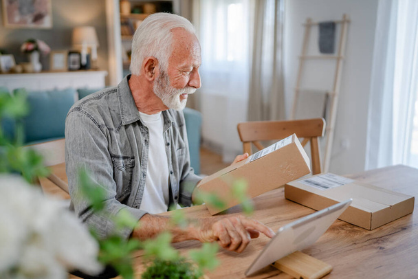 Jeden starszy mężczyzna kaukaski w domu hold cardbox pakiet dostarczone prezent lub zamówienie użyć tabletu cyfrowego online, aby sprawdzić stan zakupów przesyłki lub dla miejsca kopiowania połączeń wideo - Zdjęcie, obraz