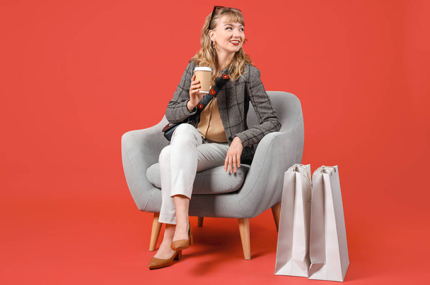 Stylish young woman with coffee cup and shopping bags in armchair on red background - Φωτογραφία, εικόνα