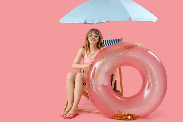 Plaj şemsiyesi ve pembe arka planda yüzme yüzüğü olan güverte sandalyesinde oturan genç bir kadın. - Fotoğraf, Görsel