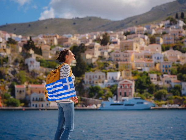 Nice asian Happy Female with backpack anf greek flag Enjoying her holidays on Symi Islands. View of port Symi or Simi, is tiny island of Dodecanese, Greece, calm atmosphere and fabulous architecture. - Photo, Image