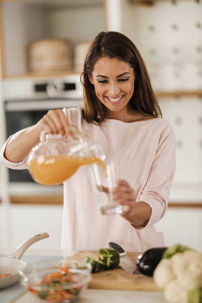 キッチンで新鮮なオレンジジュースを注いで飲んでいる若い笑顔の女性のショット. - 写真・画像
