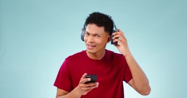 Πρόσωπο, ακρόαση και άνθρωπος με ακουστικά, χορό και streaming μουσική σε ένα μπλε φόντο στούντιο. Πορτρέτο, πρόσωπο ή χαρούμενο μοντέλο με ακουστικά, ακοή ή ήχο με smartphone, ψηφιακή εφαρμογή ή ενέργεια. - Πλάνα, βίντεο
