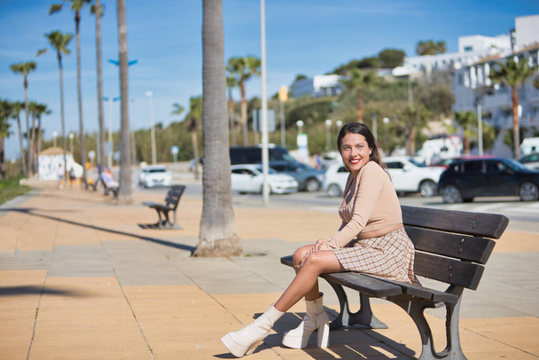 Mujer joven y atractiva, con ojos azules, suéter, falda a cuadros y plataformas blancas, sentada en un banco, feliz y sonriente. Concepto felicidad, sonrisas, miradas. - Foto, imagen