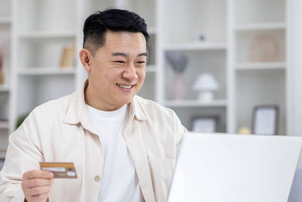 Joyful successful asian businessman working from bright home office, man using bank credit card and laptop for online shopping in online store, and booking services remotely. - Photo, Image