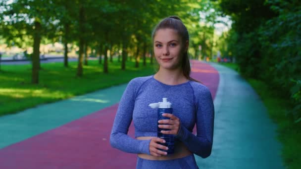Retrato de una joven mujer caucásica sosteniendo una botella llena de agua antes de correr en el parque. Fotografía con cámara de helio RED en 8K.   - Metraje, vídeo