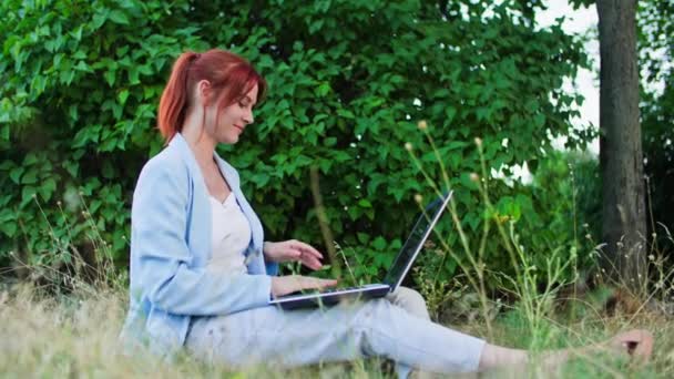 na volné noze, mladá kancelářská pracovnice pracující s laptopem, zatímco sedí na travnatém pozadí stromů v parku - Záběry, video