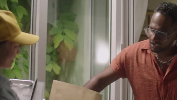 Junger schwarzer Mann öffnet Tür, nimmt Paket von Kurierin, spricht mit ihr und unterschreibt Lieferschein, während er Online-Bestellung zu Hause erhält - Filmmaterial, Video