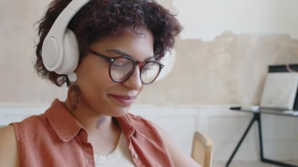 Primer plano inclinación abajo tiro de la cara y las manos de la joven mujer afroamericana en gafas y auriculares inalámbricos sentados en casa, balanceo cabeza a la música, riendo y escribiendo en el teléfono inteligente - Imágenes, Vídeo