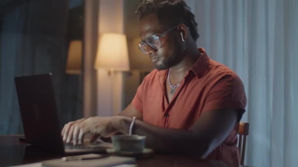 Keskivaikea laukaus musta mies langattomissa kuulokkeet kirjoittamalla kannettavan tietokoneen ja juominen teetä työskennellessään verkossa kotoa yöllä pimeässä olohuoneessa - Materiaali, video