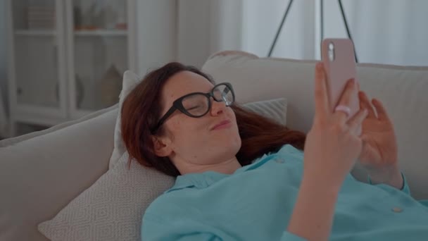 młoda kobieta z rudymi włosami leży na kanapie trzymając smartfona przewijanie wiadomości paszy wpisując komentarze - Materiał filmowy, wideo