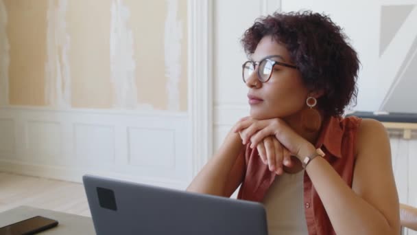 Retrato de primer plano mediano de una joven afroamericana reflexiva con cabello corto ondulado, con gafas sentadas en la mesa con computadora portátil, barbilla en las manos, mirando a la ventana y luego girando hacia la cámara - Metraje, vídeo