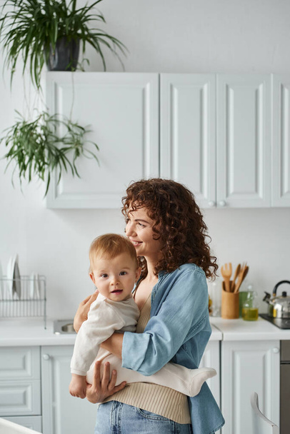 joyful woman with wavy hair embracing toddler daughter in kitchen at home, love and tenderness - Photo, image