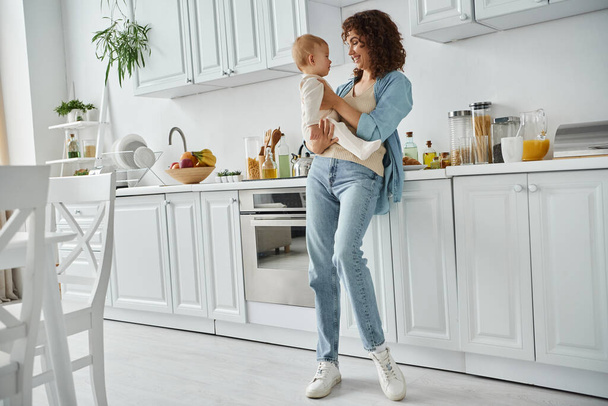 full length of happy woman holding little daughter in spacious modern kitchen with white furniture - Foto, imagen