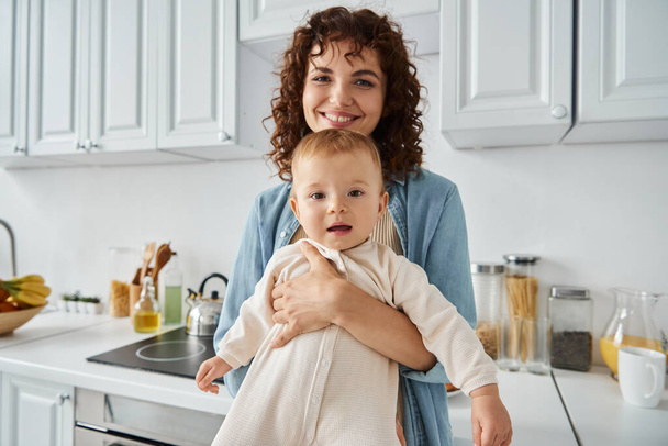 kochająca matka z falowanymi włosami przytulająca uroczą córkę w romper w domu w kuchni, rodzinna błogość - Zdjęcie, obraz