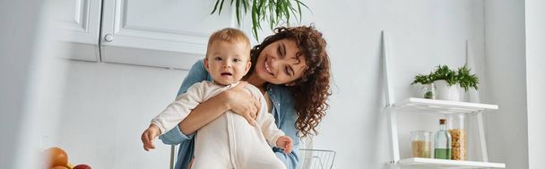 radostná žena s vlnitými vlasy objímající malé dítě v kuchyni doma, šťastné mateřství, prapor - Fotografie, Obrázek