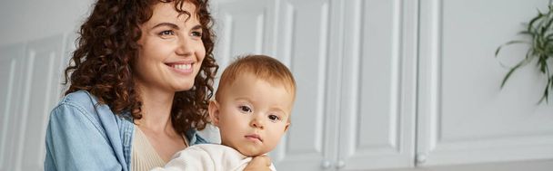 happy woman with wavy hair smiling and looking away near adorable baby girl in kitchen, banner - Photo, Image