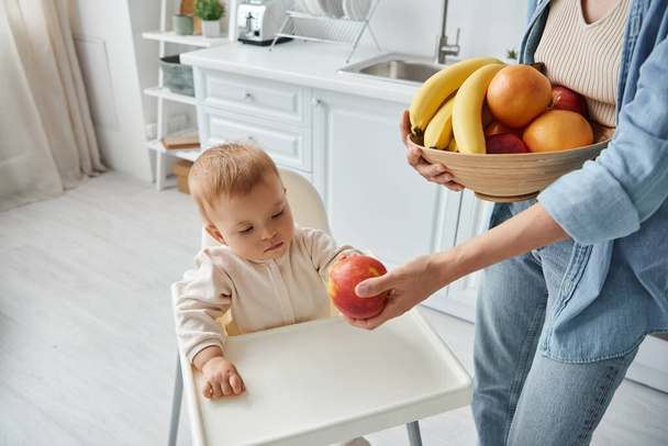 Mutter mit Schale mit frischem Obst schlägt ihrer kleinen Tochter im Kinderstuhl einen reifen Apfel vor - Foto, Bild