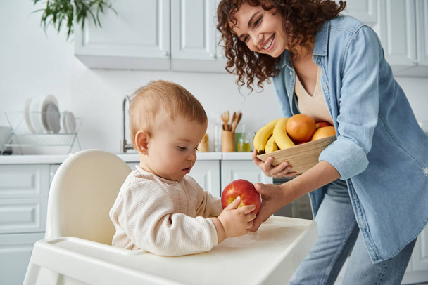 cheerful woman giving ripe apple to toddler daughter sitting in baby chair in kitchen, breakfast - Photo, Image