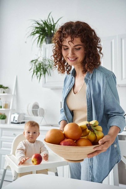 glückliche Frau mit Schale mit reifen Früchten in der Nähe von Kleinkind Tochter sitzt im Babystuhl in der Morgenküche - Foto, Bild