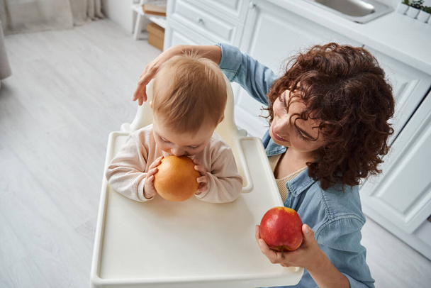 熟したリンゴと笑顔の母親の近くにオレンジを噛む赤ちゃんの椅子の小さな子供のトップビュー - 写真・画像