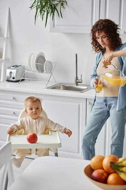Mutter gießt natürlichen Orangensaft neben Kind im Babystuhl und frisches Obst in moderne Küche - Foto, Bild