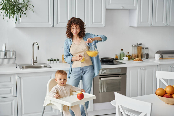 cheerful woman pouring orange juice next to little daughter in baby chair with ripe apple, breakfast - Фото, зображення
