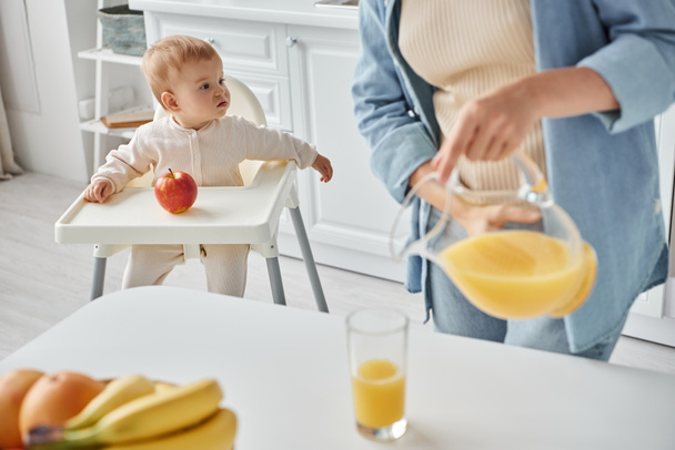 женщина наливает свежий апельсиновый сок в то время как дочка сидит в детском кресле возле спелого яблока - Фото, изображение