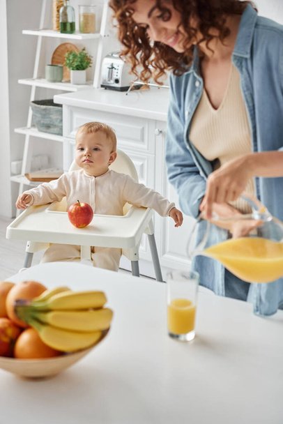 χαριτωμένο παιδί κάθεται σε καρέκλα μωρό κοντά στο μήλο, ενώ ευτυχισμένη μαμά ρίχνει φρέσκο χυμό πορτοκάλι για πρωινό - Φωτογραφία, εικόνα