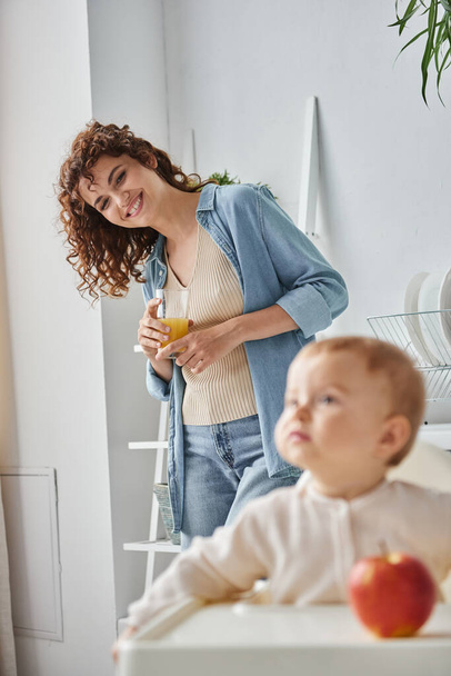 Lächelnde Mutter mit einem Glas Orangensaft und Blick auf Kleinkind-Tochter im Babystuhl neben reifem Apfel - Foto, Bild