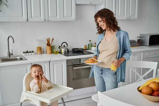 smiling woman with croissant and orange juice looking at funny child chewing wooden tongs in kitchen - Foto, Imagem