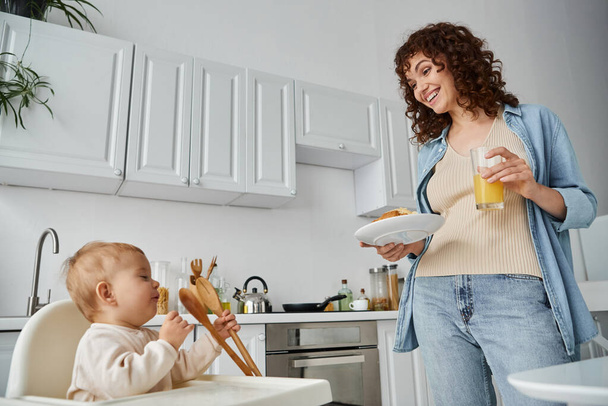 веселая женщина с круассаном и апельсиновым соком рядом с маленькой девочкой играет с деревянными щипцами на кухне - Фото, изображение