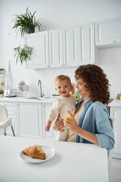 mujer sonriente sosteniendo bebé feliz y vaso de jugo de naranja fresco durante el desayuno en la cocina moderna - Foto, Imagen