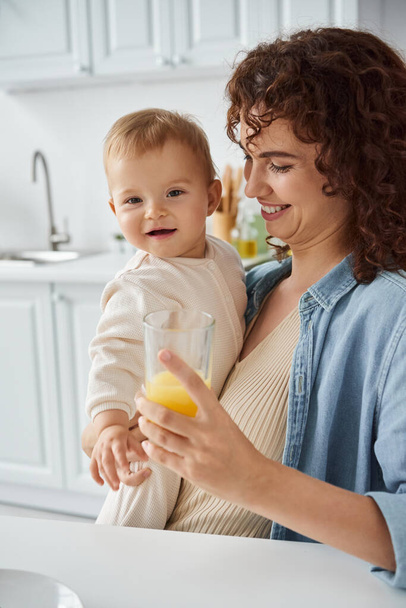 smiling woman holding glass of natural orange juice near carefree baby girl, happy family morning - Photo, Image