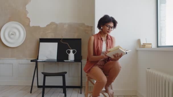 Plan complet moyen de la jeune afro-américaine en vêtements décontractés assis sur un tabouret par la fenêtre dans le salon branché de l'élégant appartement minimaliste, livre de lecture, et en regardant la caméra avec un sourire confiant - Séquence, vidéo