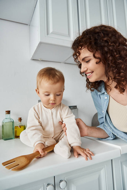 cheerful woman near little daughter with wooden fork sitting on kitchen worktop, happy morning - Foto, imagen