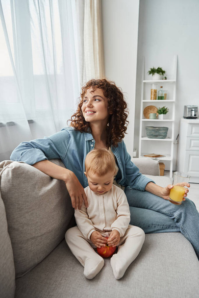 entzückte Frau mit Orangensaft schaut auf Couch neben entzückendem Kind mit Apfel weg, Glück - Foto, Bild
