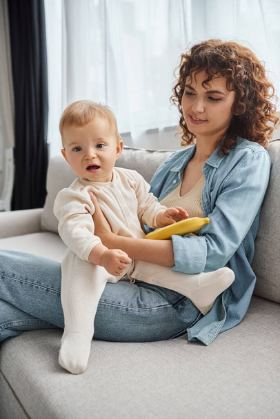 curly woman and baby girl with fresh banana sitting on cozy couch in living room, leisure time - Photo, Image