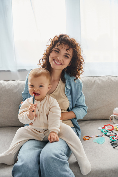 curly woman smiling at camera near toddler daughter chewing teething toy on couch in living room - Photo, Image