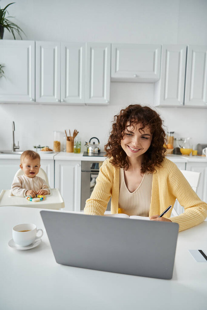 χαμογελαστή γυναίκα που εργάζεται σε φορητό υπολογιστή κοντά στην κούπα του καφέ και την κόρη νήπιο στο παρασκήνιο στην κουζίνα - Φωτογραφία, εικόνα