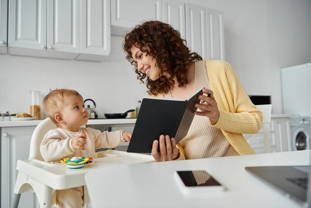 cheerful woman showing notebook to little daughter sitting in baby chair in kitchen, working parent - Photo, Image