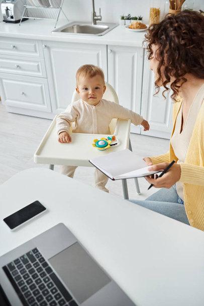 милый ребенок в детском кресле рядом мама работает с ноутбуком и ноутбуком на кухне, баланс трудовой жизни - Фото, изображение