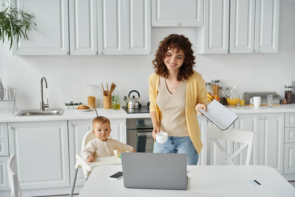 Dizüstü bilgisayarının yanında not defteri ve kahve fincanı olan mutlu bir kadın ve mutfakta bebek koltuğunda oturan küçük bir kız. - Fotoğraf, Görsel