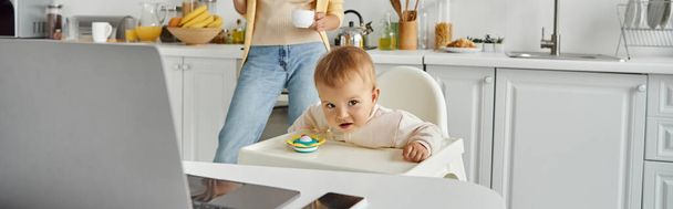 little girl sitting in baby chair in kitchen near mom with coffee cup and laptop on table, banner - Photo, Image