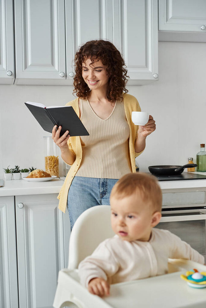 smiling woman with coffee cup looking in notebook near toddler daughter in baby chair, multitasking - Photo, Image
