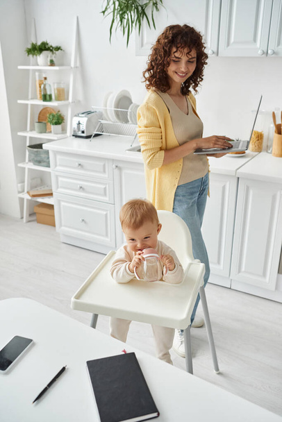 lindo niño masticando biberón cerca de la madre con computadora portátil y portátil con teléfono inteligente en la mesa de la cocina - Foto, imagen